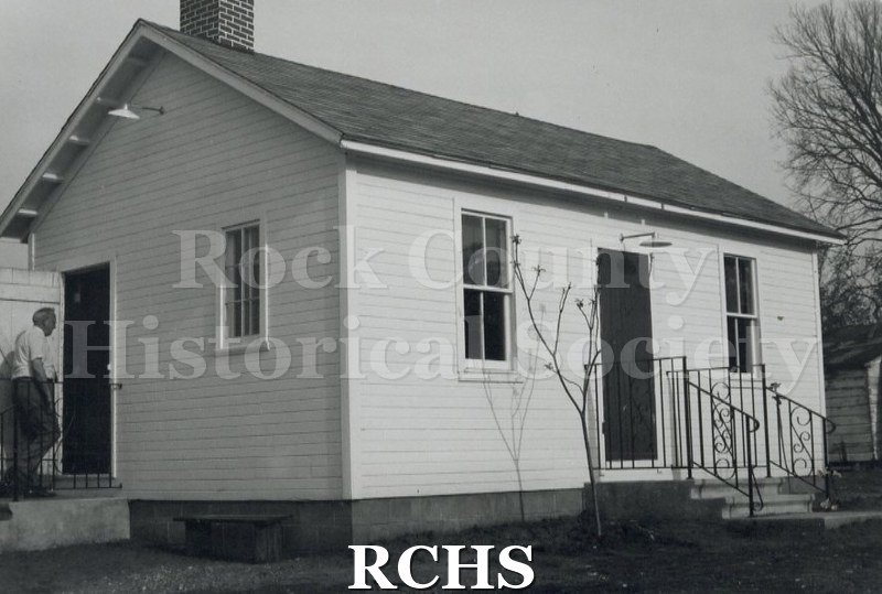 Black and white photograph of the Douglas School, a one room school house. There is a man entering the door on the left.