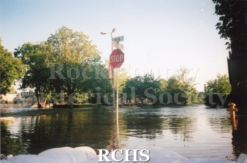Color photo of the 2008 flood in Janesville. In the center is a stop sign with the street signs for Main St. and Pease Ct. Water floods the intersection.