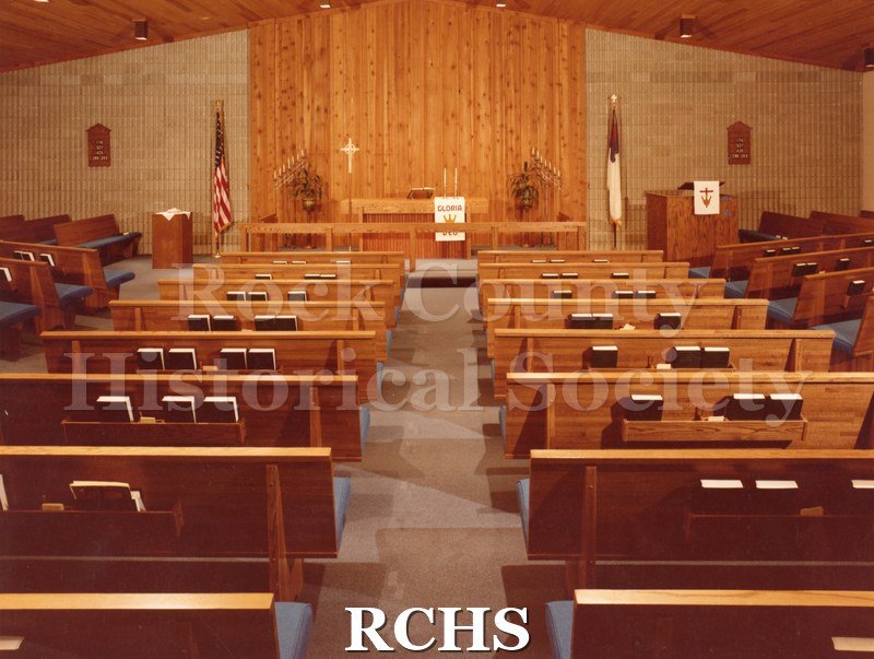 Colored photograph of the interior of an unidentified church in Neenah, WI, looking from the back to the altar. The altar has a white parament with "Gloria" on it.