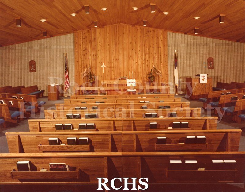 Colored photograph of the interior of an unidentified church in Neenah, WI, looking from the back to the altar. The altar has a white parament with "Gloria" on it.