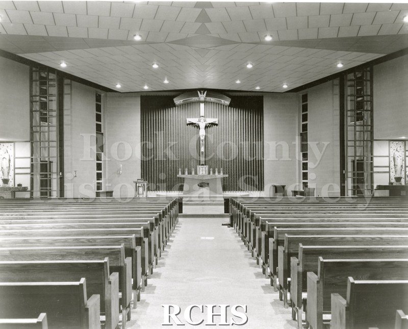 IPhotograph. Black and white photograph of the interior of an unidentified church showing the pews and altar area. There is a large crucifix over the altar.
