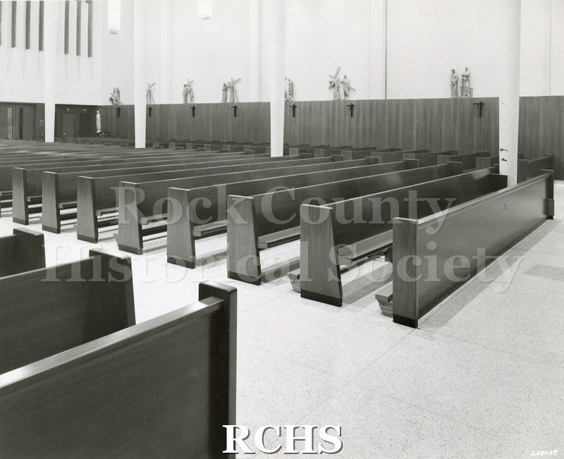 Black and white photograph of the interior of an unidentified church showing the pews from the side. There are stations of the cross on the back wall.