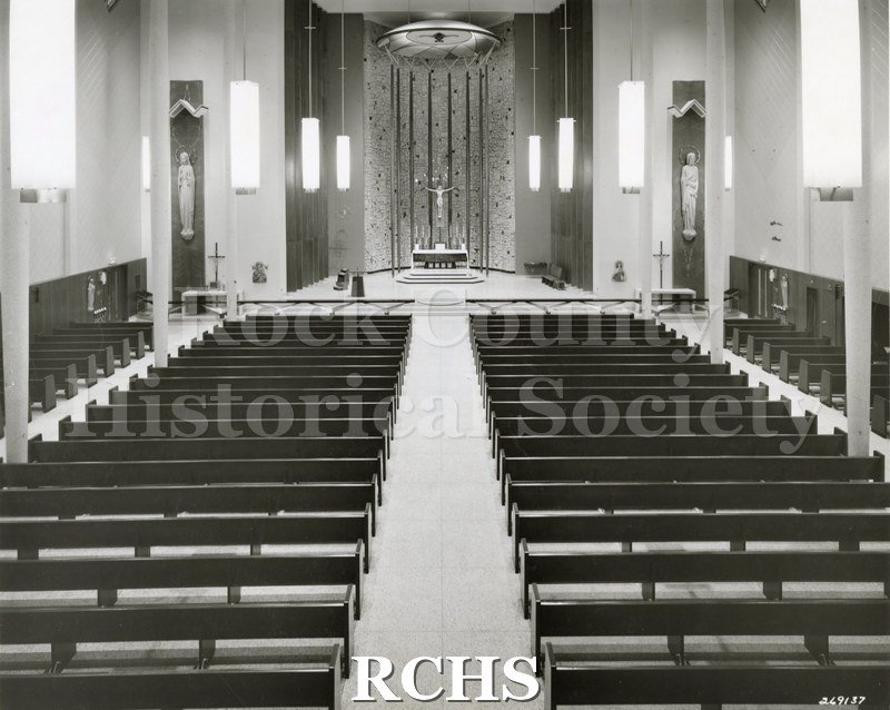 Black and white photograph of the interior of an unidentified church showing the pews leading up to an altar with a crucifix on it.