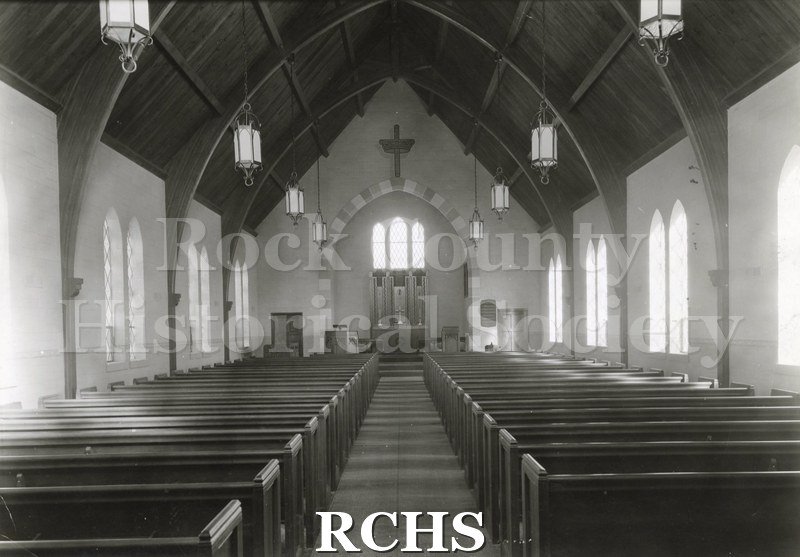 Black and white photograph of the interior of Lutheran Church in Saginaw, MI., featuring the pews, an altar in a curved opening with a large cross on top of the.