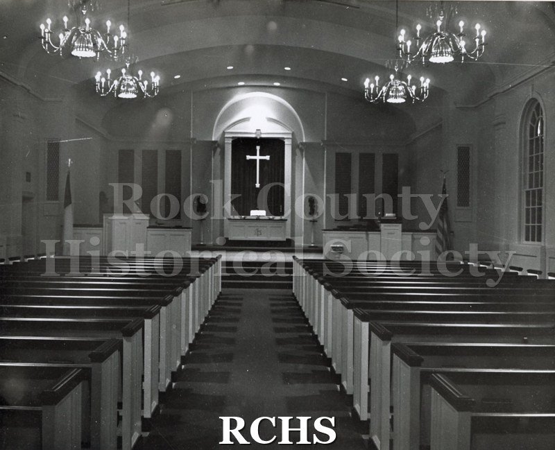 Black and white photograph of the interior of an unidentified church in Clear Lake, IA, looking toward the front. There is an illuminated cross above the altar.