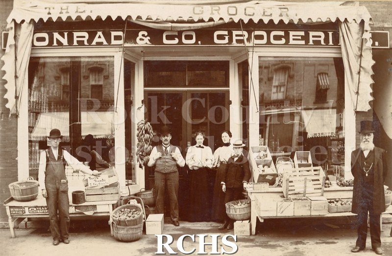 Black and white photograph of exterior of "Conrad & Co, Groceries." 2 women, 3 men, and 1 boy visible.