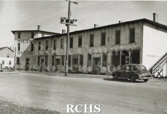 Black and white photograph of the Milton House. Exterior appears dirty. Telephone pole in center with wires stretching out to both sides.