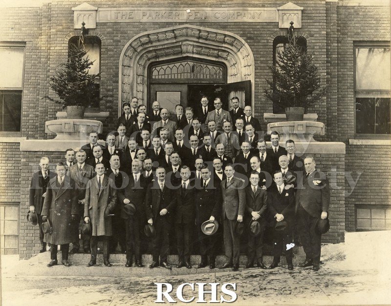 Black and white photograph of male Parker Pen employees in suits and ties standing outside of the company building's entrance. There is snow on the ground.
