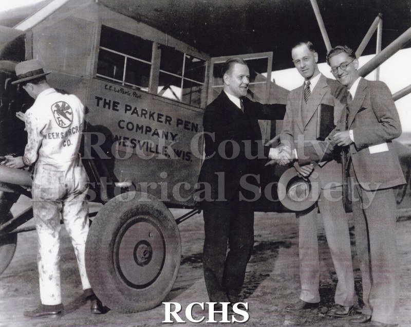 Black and white photograph of four men are next to a plane that reads "The Parker Pen Company/Janesville, Wis." The photograph is accompanied by a white piece of.