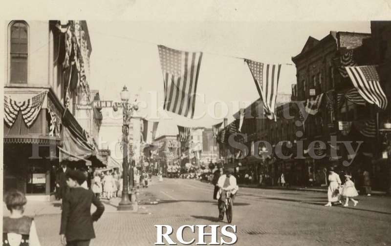 Historic image of Downtown Janesville on 4th of July. There are American flags above the street and people on the sidewalks and in the road.