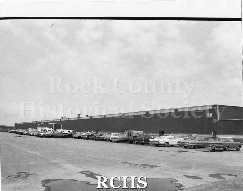 Black and white photograph of Highway Trailer Co., Edgerton, WI. Shows long building with an H on the right side and a line of cars parked in front of the building.