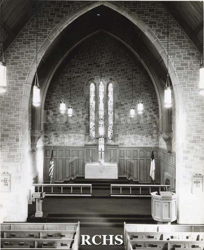 Black and white photograph of the interior of an unidentified church, showing the altar area. There are three narrow stained glass windows above the altar.
