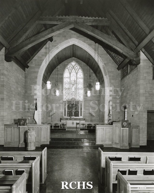 Black and white photograph of the interior of an unidentified church, showing the altar with an arched stained glass window behind it.