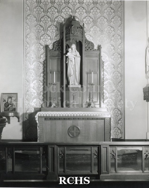 Black and white photograph of the interior of an unidentified church with a wooden altar. Above the altar is a statue of the Virgin Mary and child.