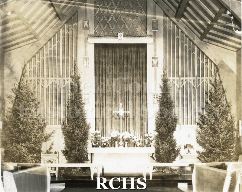 Black and white photograph of the interior of an unidentified church at Christmas time with evergreen trees decorating the chancel.