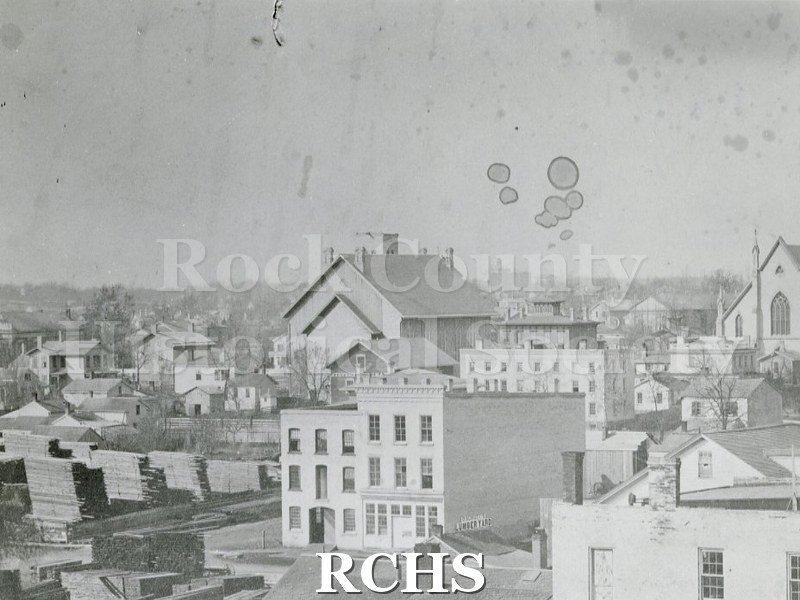 Black and white photograph of early Janesville. There is a three story building in the foreground with piles of lumber beside it and other buildings in the.