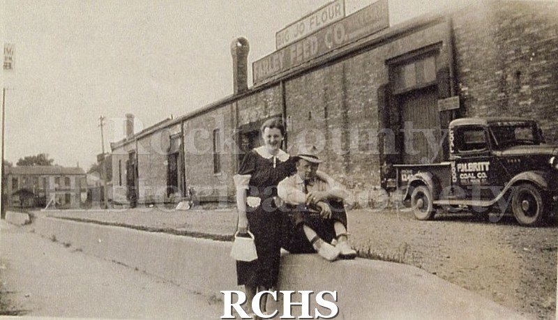 Black and white photograph of an unidentified man sitting on a cement wall and a woman standing beside him. They are in front of Farley Feed Co.