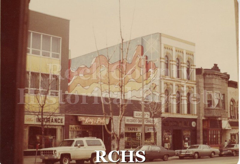 Colored photograph of a three story building with a mural on the left side on Main Street. Beside it is the King's Pub and Dorothy's Records.
