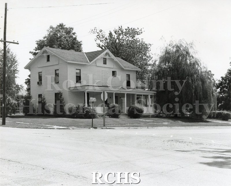 Black and white photograph of an unidentified two story sided house with a porch extending along the length of the front.