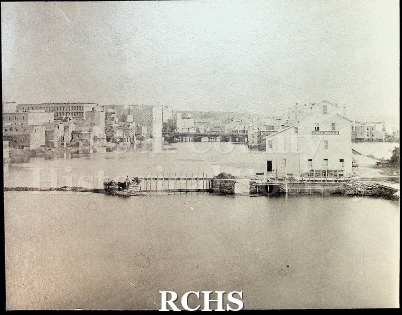 Historic image of Black and white copy print (1984) of early Janesville, showing a two story building on the river on the right side.