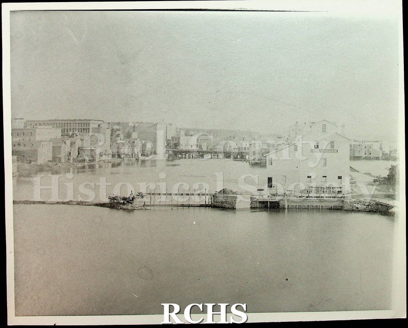 Black and white photograph of early Janesville, showing a two story building on the river on the right side.