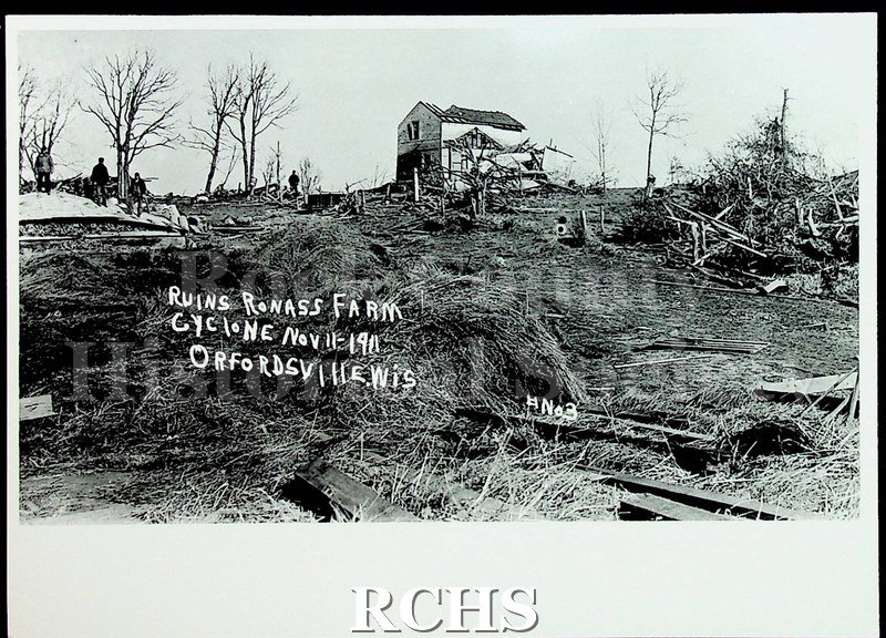Black and white photograph of the aftermath of a cyclone. Shows the debris field and a house with its roof torn off. People are pictured in the background.