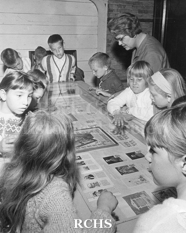 Black and white photograph of school children on a field trip. Shows schoolchildren looking at newspapers under glass at the carriage house on the Tallman campus.