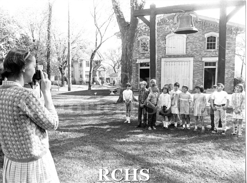 Black and white photograph of schoolchildren on a field trip. They are standing in front of the carriage house with a large bell on a wooden mounting.