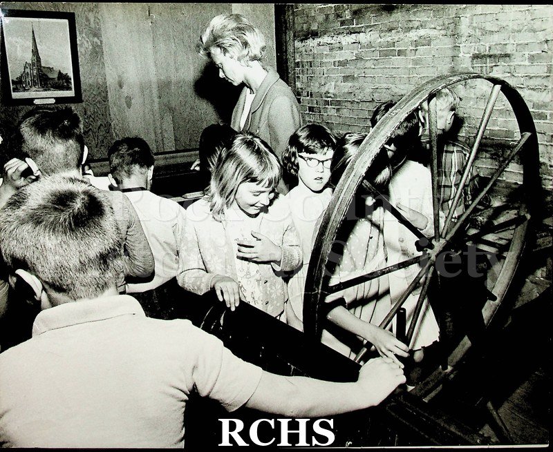Black and white photograph of school children on a field trip. Shows schoolchildren looking at a wheel and other exhibits at the carriage house.