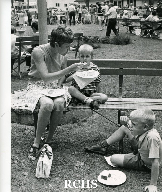 Black and white photograph of the 1972 Arts & Crafts Fair. A mother and her two sons are enjoying cake and ice cream.