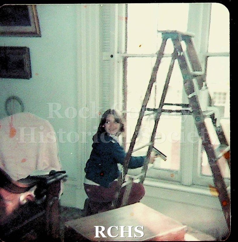 Color photo of the Opening Tallman House, April 1977. Shows a young woman kneeling beside a ladder.