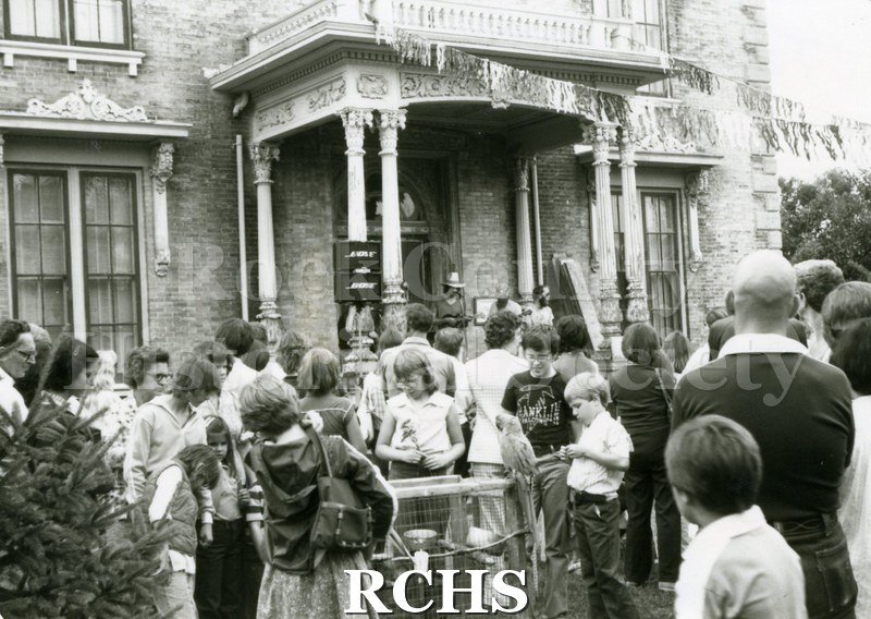 Black and white photograph of many people standing on the lawn in front of the Tallman House. Some are looking at a display.