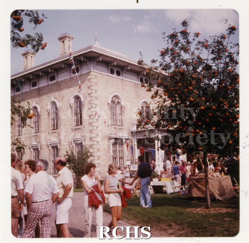 Color photo of Tallman Arts Festival. There are several people standing on the grounds. Tallman House is visible in the background.