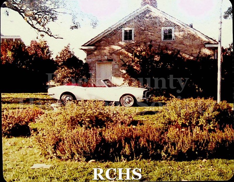 Color photo of a white convertible with the top down in front of the Stone House.