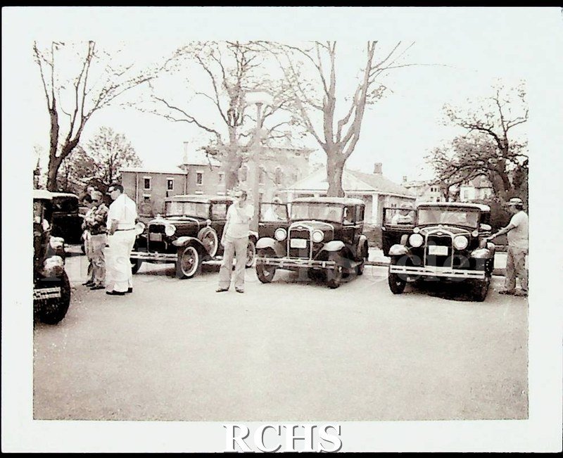 Black and white photograph of Model A Fords parked in front of the Tallman House. There are people standing beside the cars.