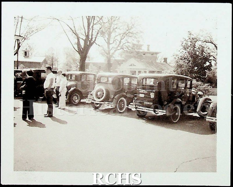 Black and white photograph of several Model A Ford cars parked in front of the Stone House, shown from the rear. There are people standing around them.