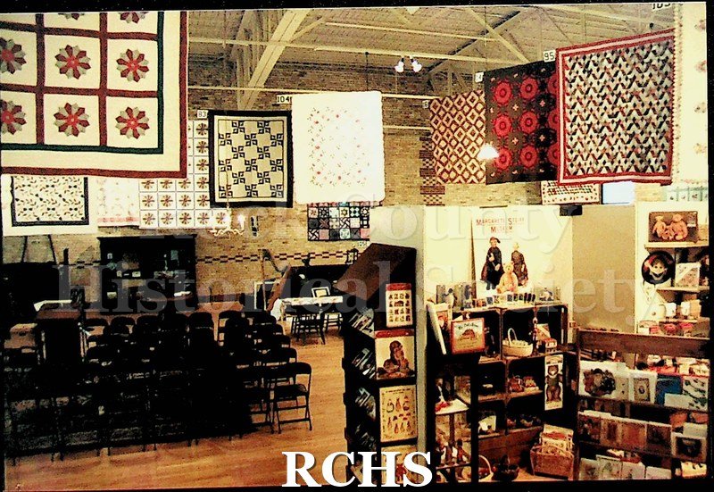 Colored photograph of several quilts hanging on display at the Tallman House museum. There are chairs set up in front of them.
