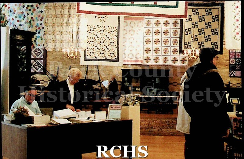 Black and white photograph of several quilts hanging on display at the Tallman House museum. There are two people at a desk in front of the photo.