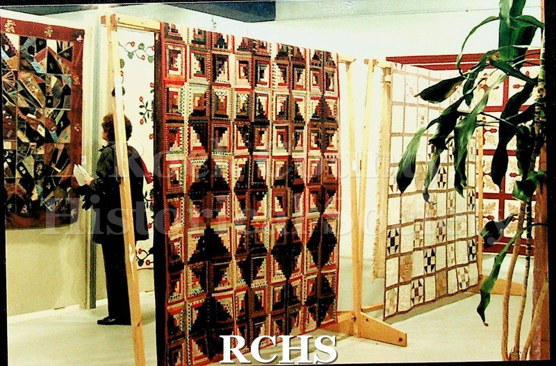 Colored photograph of several quilts hanging on display at the Tallman House museum. There is a woman standing in front of one of the quilts.