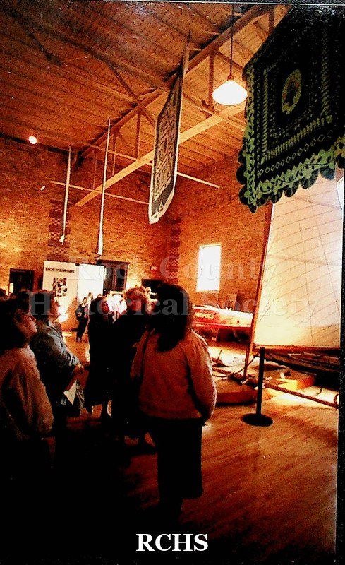 Colored photograph of several quilts hanging on display at the Tallman House museum. There are several people looking at them.