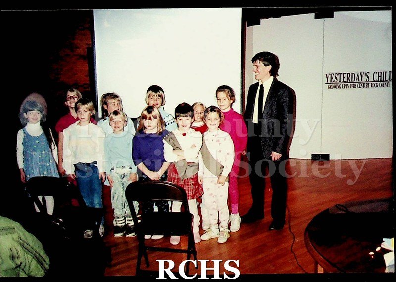 Colored photograph of several school children standing in front of a screen with Mike Brubaker standing beside them.