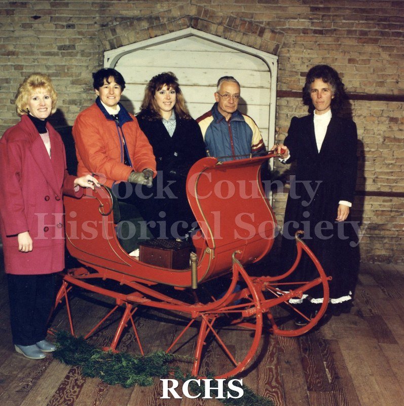 Colored photograph of Dana Thorpe, Ann Wilmot, Dee Locke, Maurice Montgomery and Mary Ann Ambrose standing by a sleigh in the basement of the Museum.