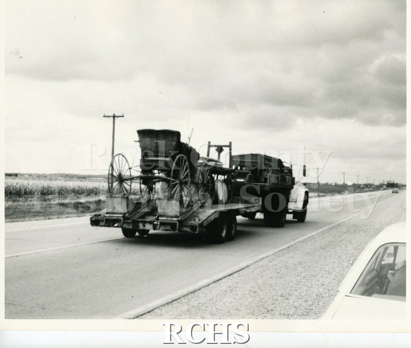 Black and white photograph of a sleigh on the back of a truck going down the highway.
