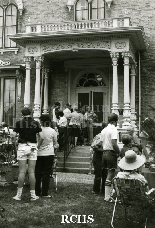 Black and white photograph of the 1983 Tallman Festival. People are standing near the Tallman House. N83013 Strip.