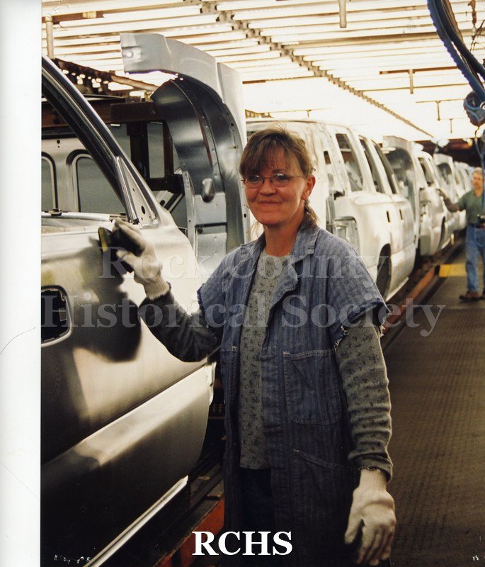 Colored photograph of an unidentified female GM worker. She is wearing glasses and white gloves, and she is at an assembly line of white cars.