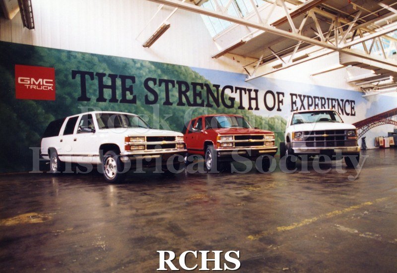Colored photograph of three GMC trucks inside a building and in front of a billboard that reads "The Strength of Experience." The first and third trucks are white.