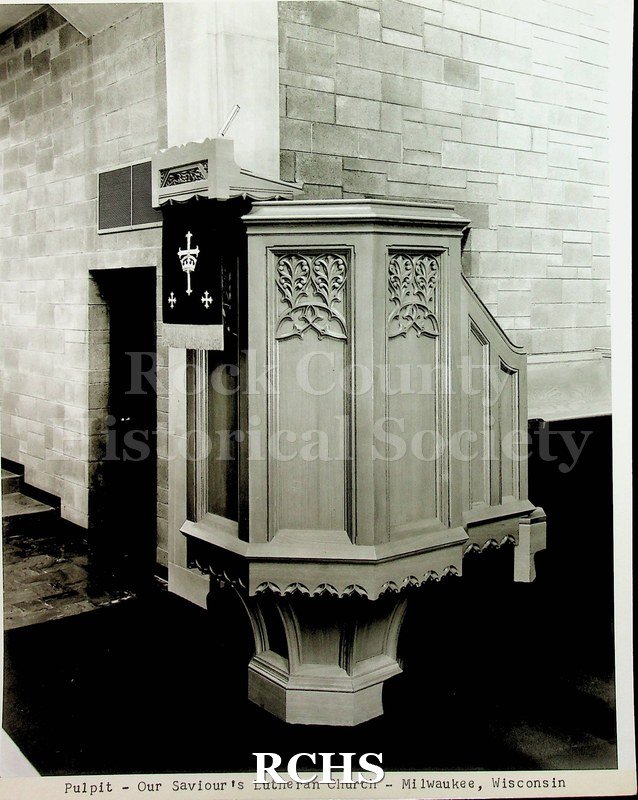 Black and white photograph of an elaborate wooden pulpit made by Ossit Furniture. Shows placement in the church.