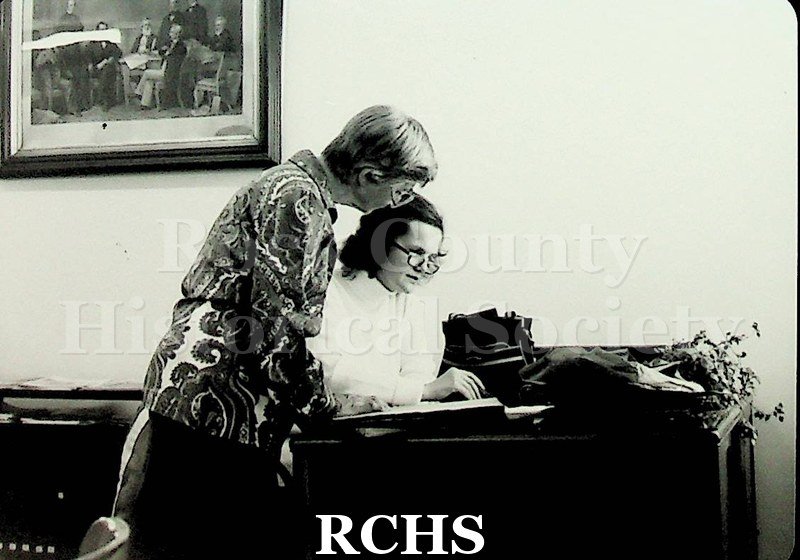 Black and white photograph of Ruth Widdicombe and a researcher. One is standing beside the other who is seated at a desk.