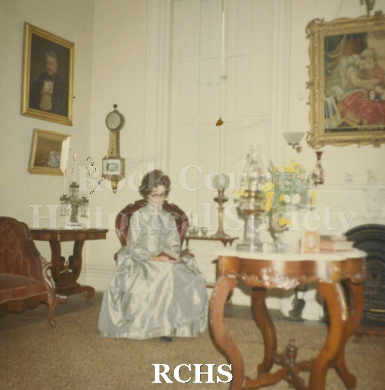 Colored photograph of an unidentified woman in costume sitting in a red chair in the Tallman House. There is a gateleg table in the front of the picture.