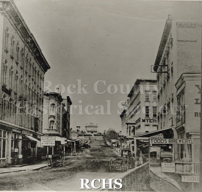 Black and white photograph of Early Janesville. Shows several buildings on the right side, including Myers and Todd's Brewery. On the left is a sign for "H. A.